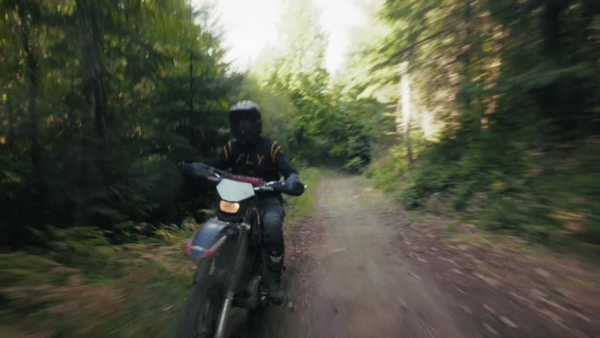 The rider wears dark protective gear and a helmet while navigating a red and black dirt bike across the uneven wooded terrain