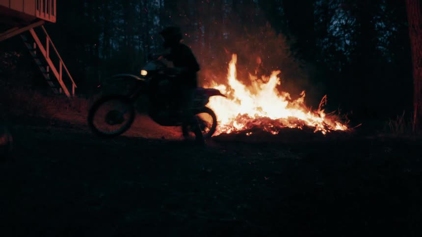 Silhouetted motorcyclist rides along a forest trail at night with fire burning in the background