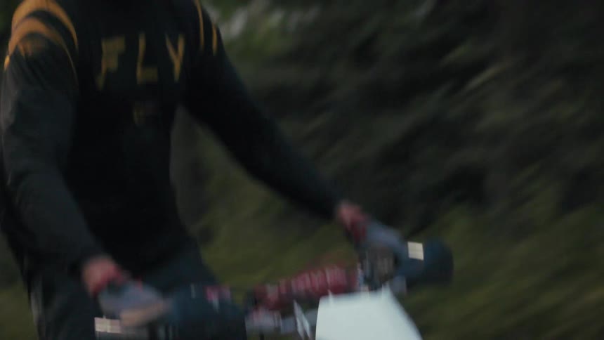A rider in a black helmet and jersey navigates a vintage honda dirt bike along a paved surfacethe camera tracks from the spinning wheels up to the riders face as they speed past a backdrop of evergreen trees