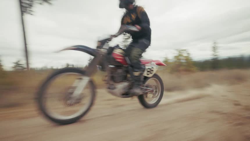 A person rides a dirt bike down a dusty gravel path through a hilly wooded landscape under an overcast sky with the camera following closely behind before panning across the autumnal terrain