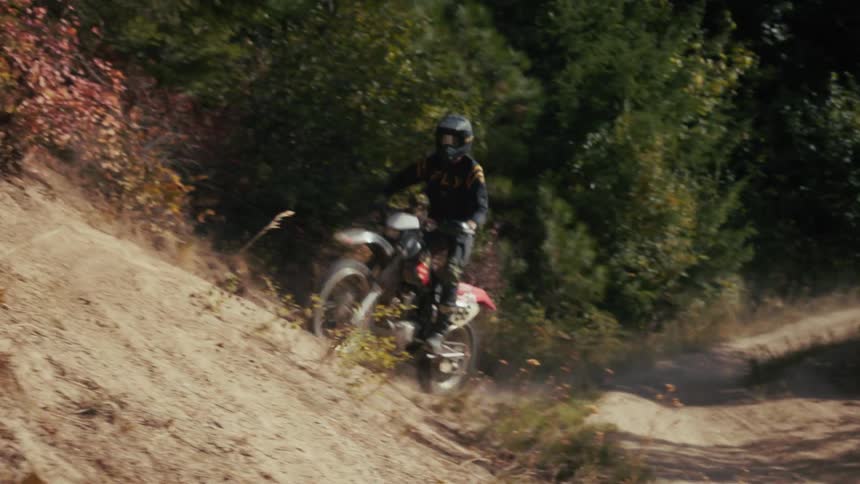 Biker wearing a helmet and dark clothes riding a motocycle on a dust road and going up a slope on an area surrounded by trees outdoors during day time