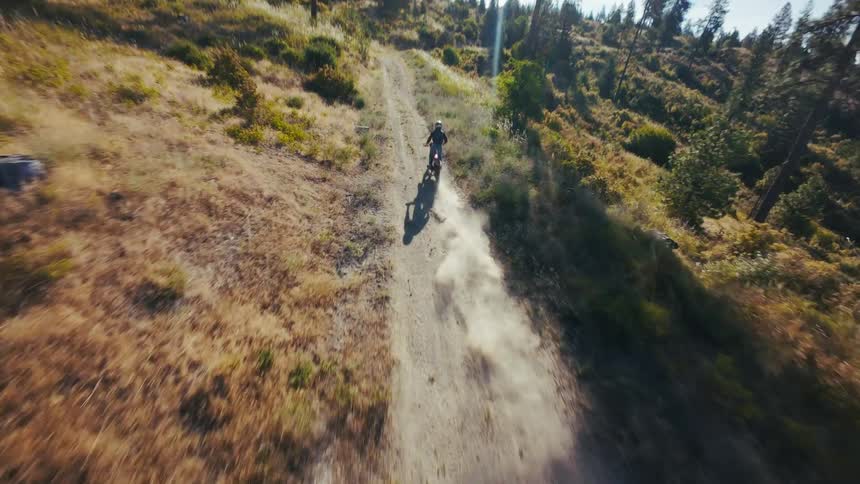 A distant view from above shows a person riding a motorcycle along a dirt road through a vast field of thin bare trees and colorful autumn brush