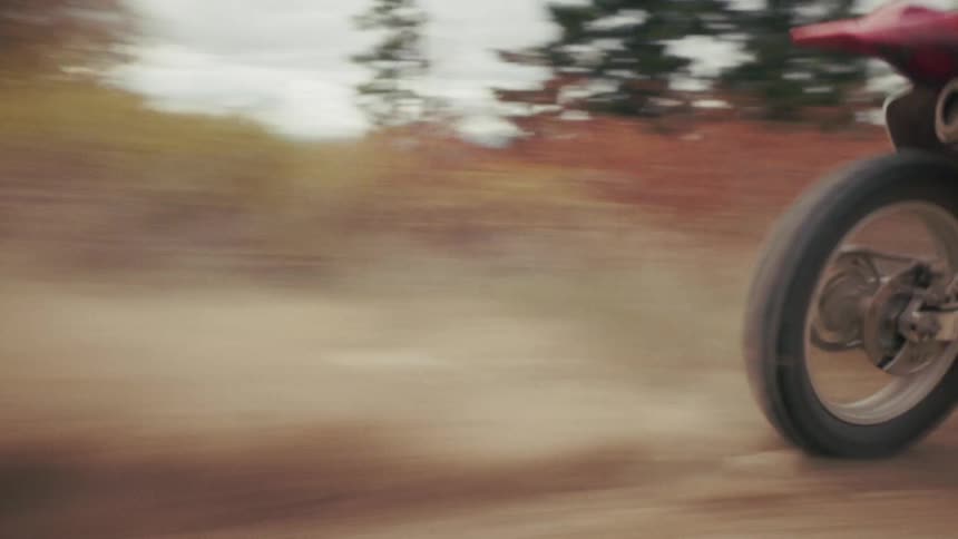 A motorcyclist speeds along a rugged dirt trail kicking up dust while navigating through a scenic autumnal forestthe camera follows at a low angle capturing the rapid movement of the wheels against the gravelly terrain as the rider disappears into th