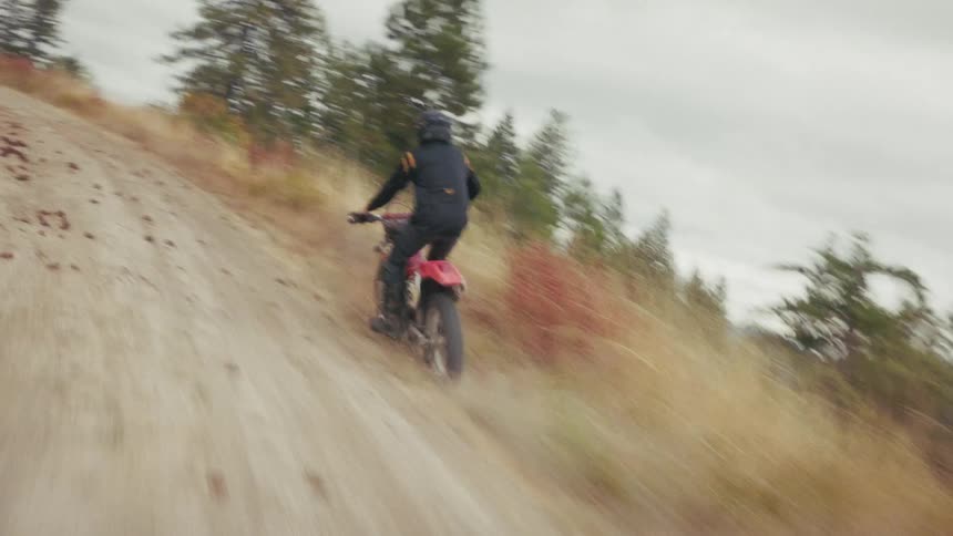 A motorcyclist wearing a black jersey and helmet rides a red dirt bike up a steep winding gravel trail through a mountainous forest under an overcast sky