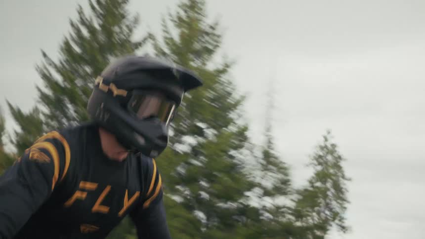 A man wearing a black helmet and a dark long sleeve jersey with gold stripes rides a motorcycle quickly on a dirt path through an open field with sparse trees under a cloudy sky
