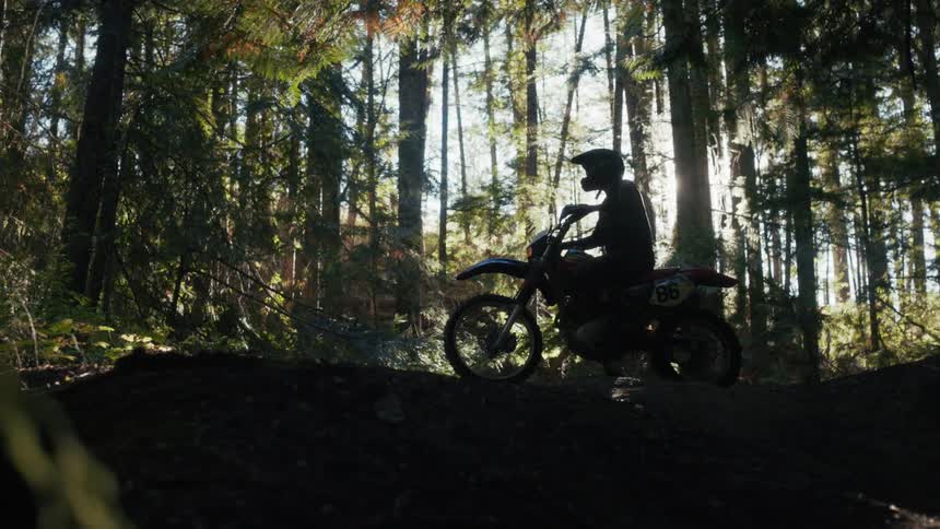 A silhouetted dirt bike rider sits motionless on a trail deep within a sundrenched dense forestglimmering light filters through the tall evergreen trees creating a dramatic backlighting effect against the dark forest floor