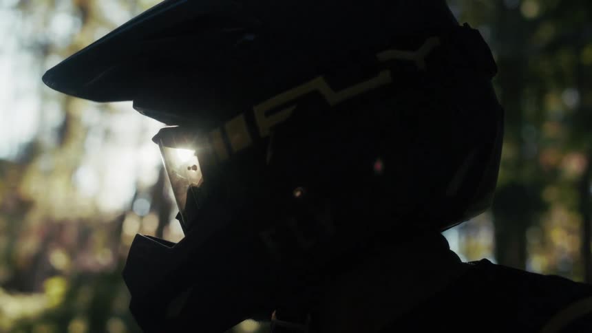 Profile of a motocross rider wearing helmet and goggles in a forest setting lit by natural light