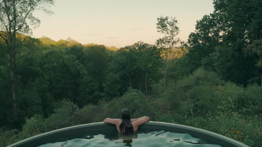 A woman with long dark hair sits in a round outdoor pool and looks out at a dense green forest during a sunset with a warm orange glow in the sky