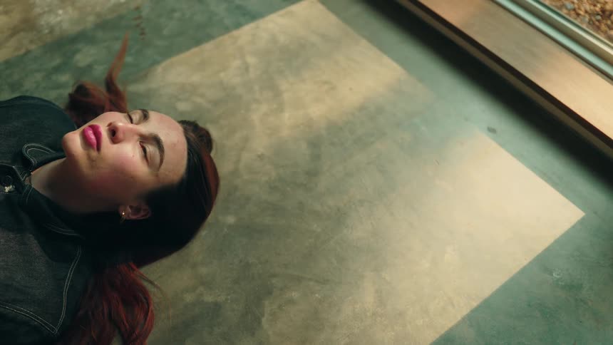 A woman gazing thoughtfully upward while lying on a concrete floor in a sunlit room