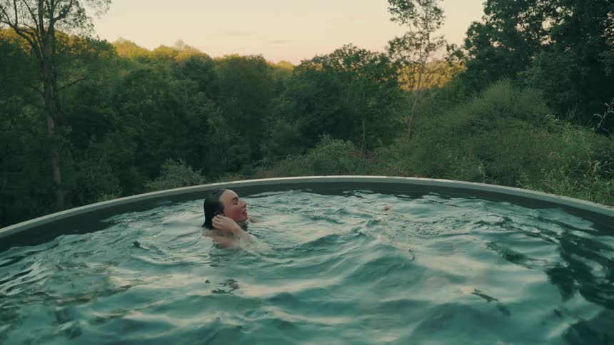 Two people laugh and splash in an outdoor pool surrounded by lush green trees enjoying the calm of the landscape at golden hour