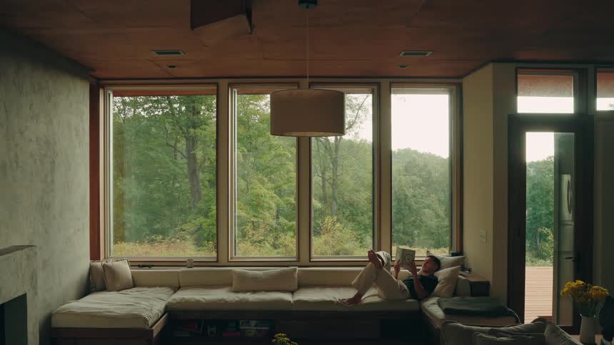Man lying on his couch reading a book with a view of forested mountains in the windows