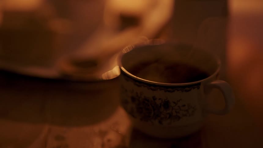 Steam rises from a floral patterned teacup filled with dark liquid sitting on a wooden surface in a warm and dimly lit room