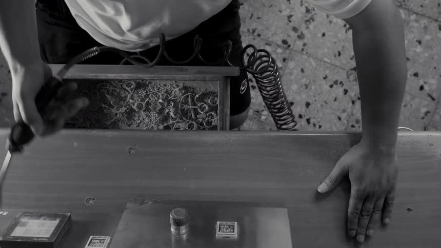 A black and white view from above shows a worker using a small handheld tool at a workbench in a workshop