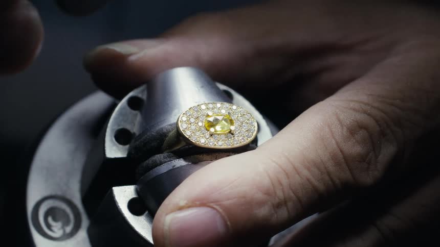 Jewelry with central yellow stone being polished using rotary tool in metal clamp