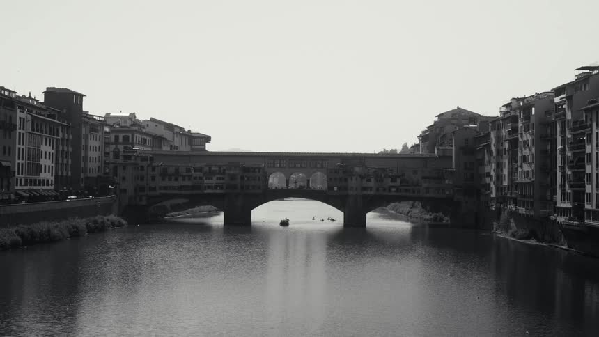 A black and white wide shot shows the ponte vecchio bridge spanning across a calm river in florence italy with historic buildings lining both sides of the water