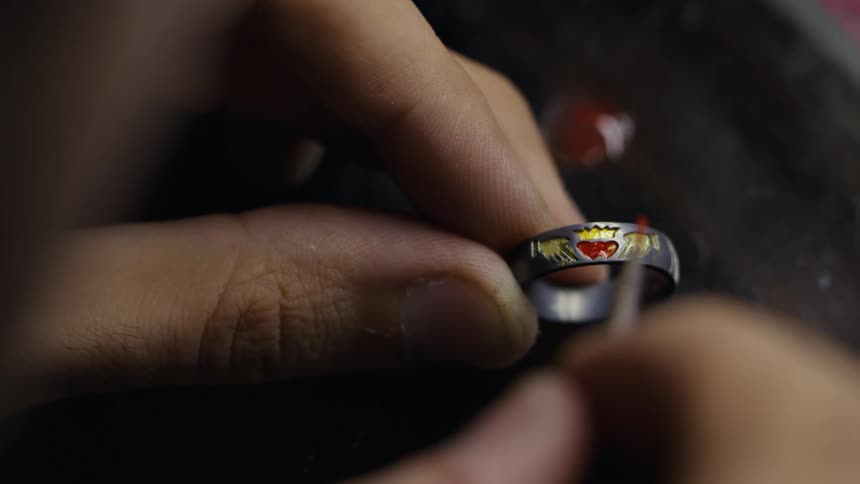 A close up shot shows a persons hands using a thin brush to carefully apply a bright red glaze to the heart shaped center of a claddagh style ring
