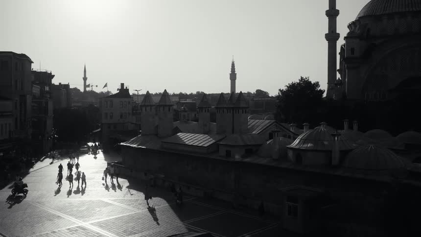 This blackandwhite aerial footage captures a busy public square in istanbul dominated by the silhouette of a large mosque and traditional domed architecture long shadows stretch across the sunlit pavement as people and a motorcycle move through the pl