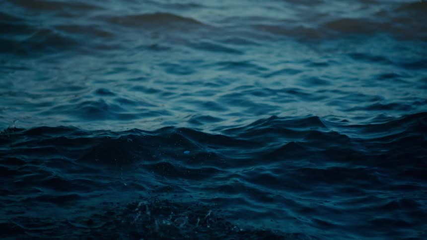 Deep blue ocean waves roll steadily across the frame under a dim lowlight sky small ripples and occasional white splashes break the surface of the dark churning water