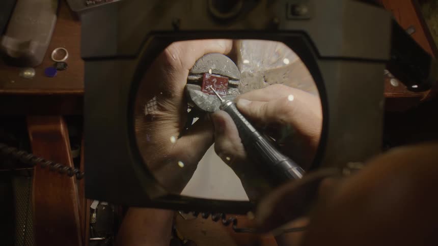 A close view through a magnifying lens shows a jeweler using a small power tool to carefully carve a detailed design into a red square gemstone held in a metal clamp