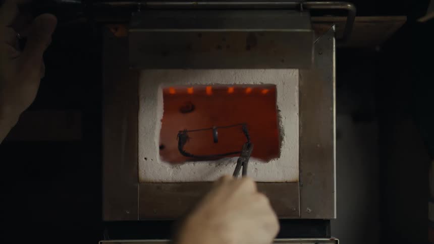 A person uses a handle to lift the door of a small industrial kiln revealing a glowing orange interior using a pair of long metal tongs they carefully reach inside to retrieve a dark heated metal component before closing the door again