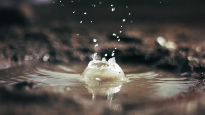 A single droplet of water falls into a shallow muddy puddle on the groundthe impact creates a small crownshaped splash and sends tiny beads of water flying upward