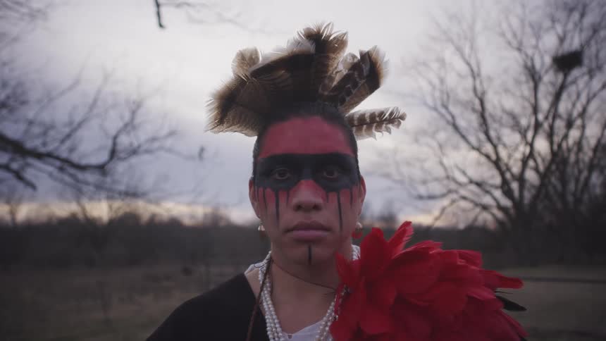 This video features a person in indigenousinspired regalia including a feathered headpiece and red and black face paint standing in a dim outdoor field they slowly turn their head to face the camera directly maintaining a steady and powerful express