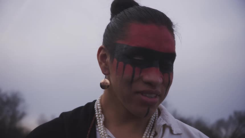 Young adult man wearing traditional native face painting and jewelry smiling with trees and the cloudy sky on the background outdoors during day time