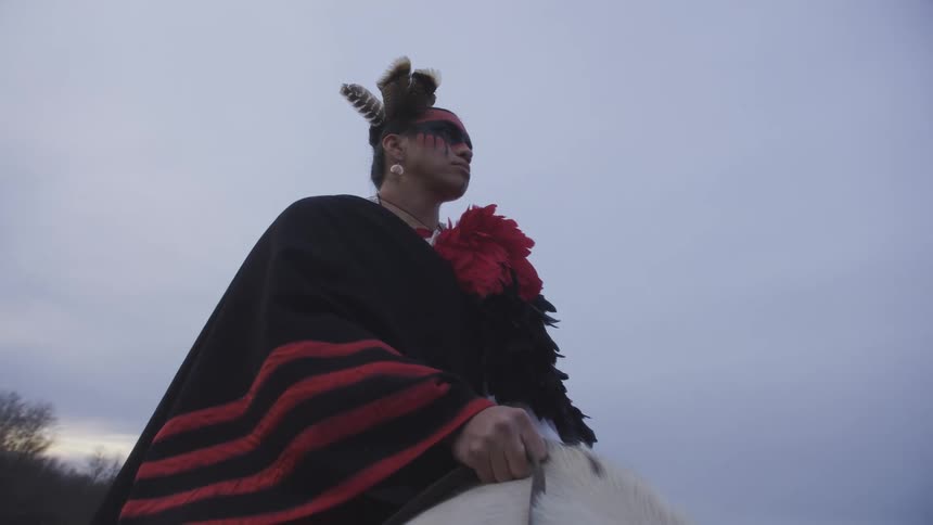 A native american person in traditional attire sits astride a horse across an open landscape at dusk