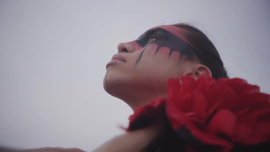 A close up profile view shows an indigenous person with red and black face paint looking upward as someones hands adjust their attire