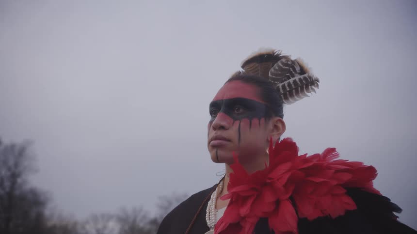 A person with traditional indigenous face paint and feathers in their hair gazes solemnly into the distance against a cloudy sky they wear red and black paint across their eyes and forehead complemented by large coppercolored earrings and bright red p