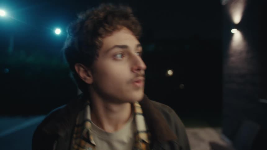 A young man with his eyes closed stands outside at night he opens them looks startled to his left and quickly walks out of the frame while a street light glows in the background