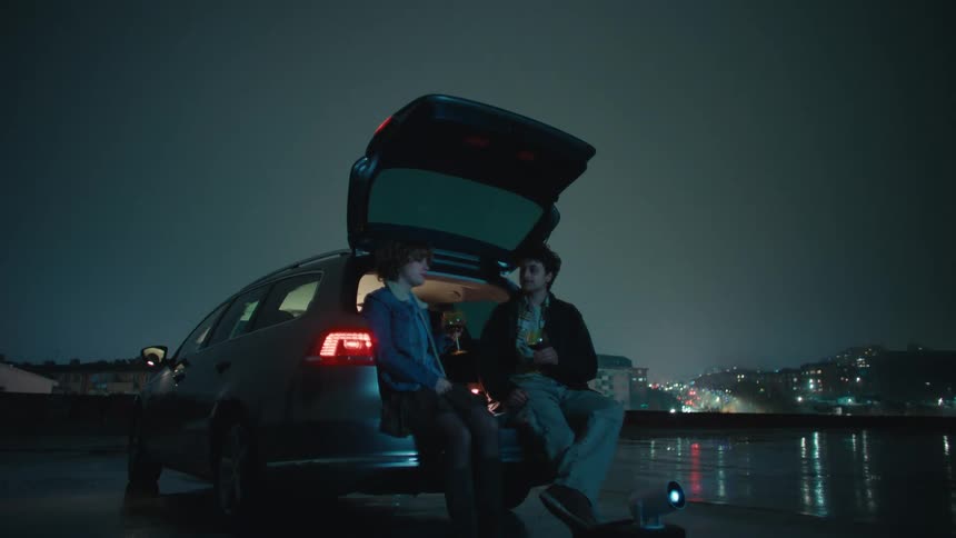 Male and female couple of teenager sitting on the open trunk of a car drinking wine and talking with a projector at their feet next to a river with a metropolitan urban neighborhood on the background outdoors during night time