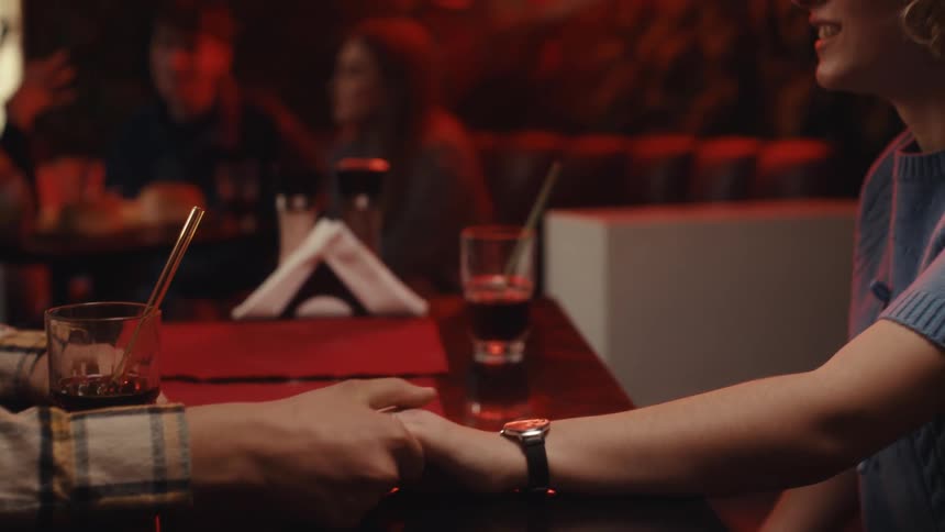 A man in a plaid shirt and a woman in a blue top sit at a table in a dimly lit bar holding hands while a neon sign glows in the background