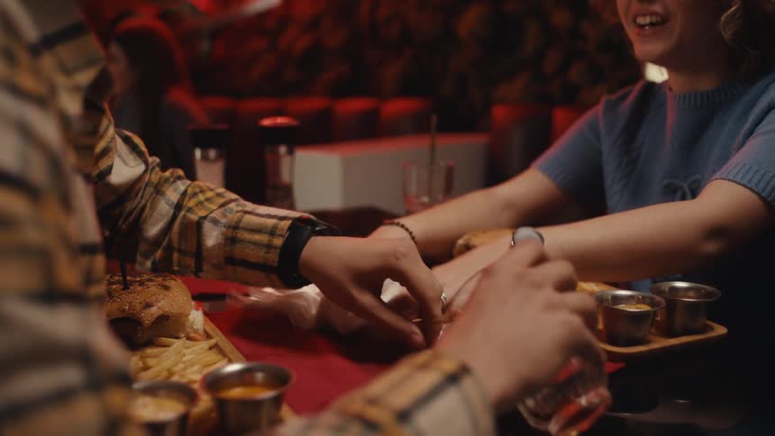 Two teenagers sitting at a table in a restaurant talking and eating as a glass spills and they try to clean it up with napkins