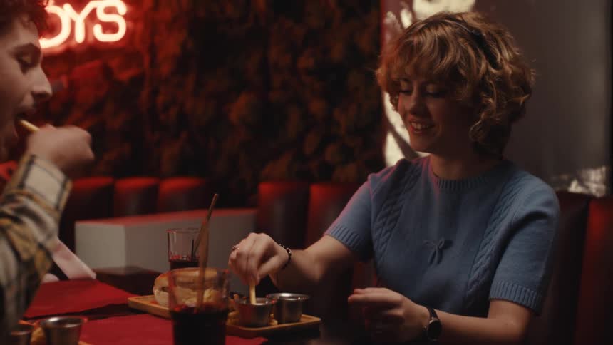 A man and a woman eat a meal together at a table in a bar while a neon sign glows in the background