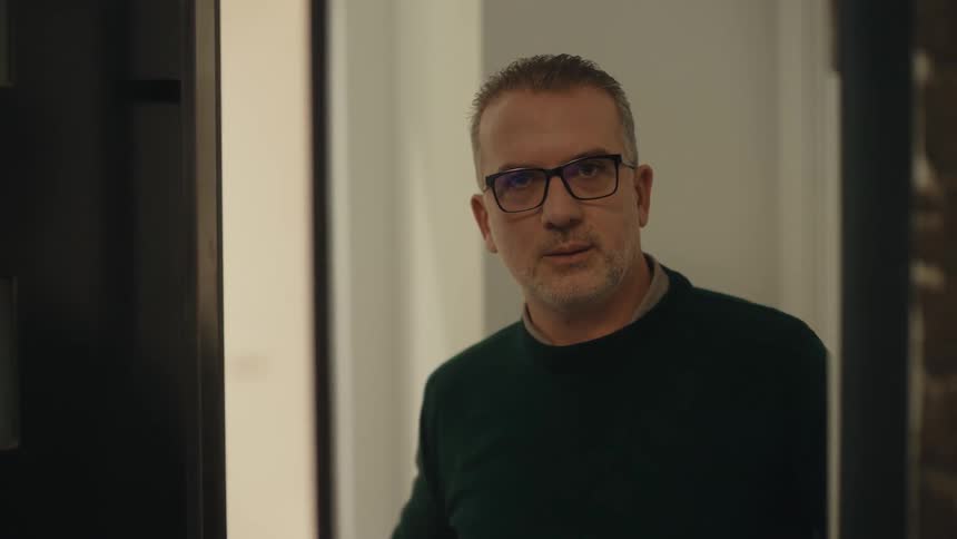 Adult man wearing glasses opening the door to a house with a nervous facial expression then closing it again after a smiling teenage girl passes by him and leaves the house outdoors during night time
