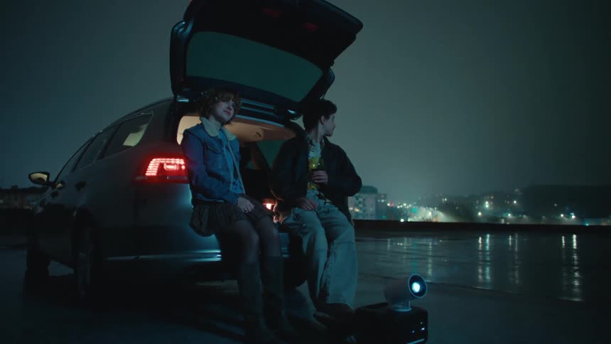 Two people sitting at the open trunk of a car in a rainy nighttime urban area with city lights in the background