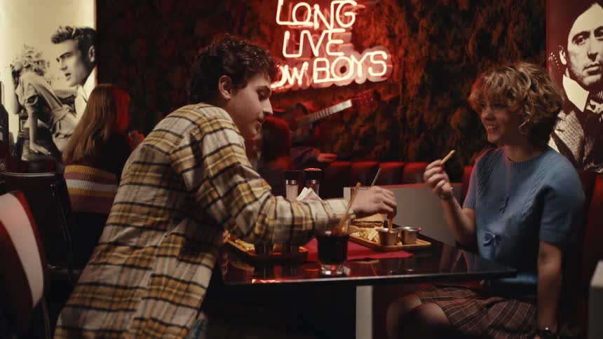 Couple seated at a table while a server places burgers in a restaurant with warm lighting and a neon sign in the background