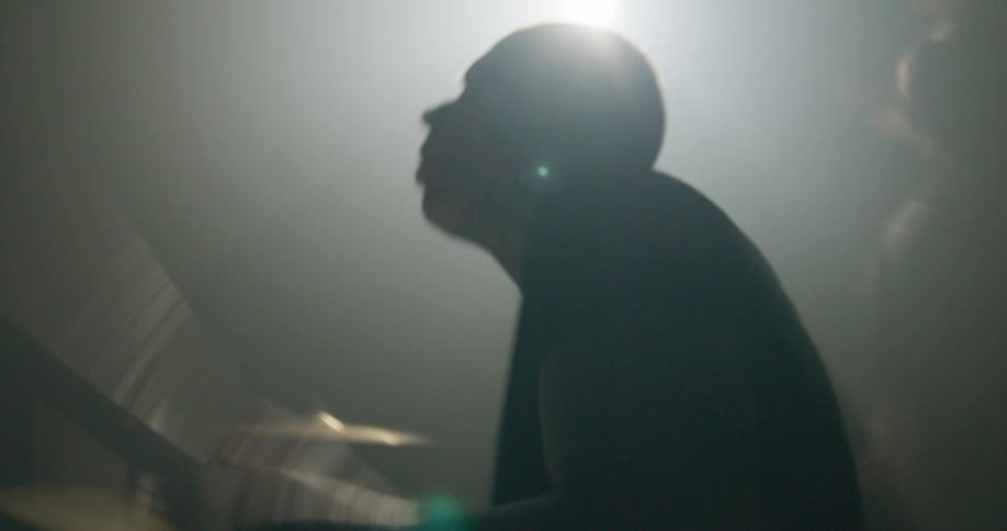 Enveloped in thick haze and blinding backlighting a silhouetted drummer strikes his instrument with raw energy while the camera captures the intense motion from behind