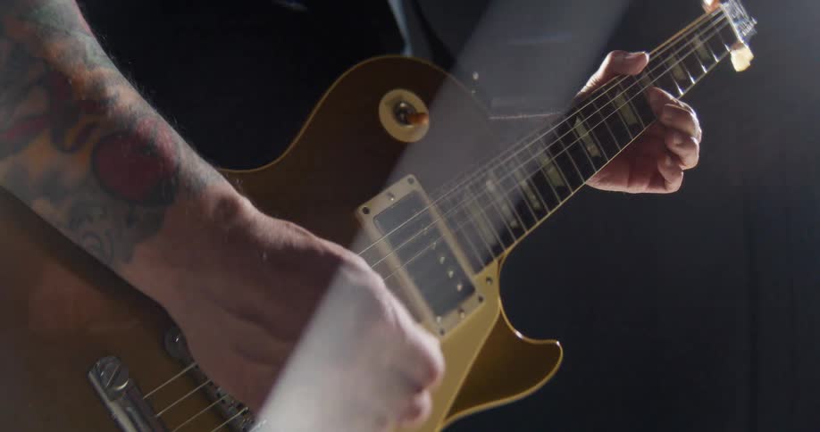 In an energetic closeup a guitarists tattooed arm anchors a gold electric guitar as their hand furiously strums the strings creating a blur of rapid motion