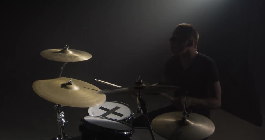 The drummer is seated behind a drum kit in a dark atmospheric studio lit by a single dramatic backlight he is captured midperformance raising his drumsticks high before striking the cymbals and snare with intense energy