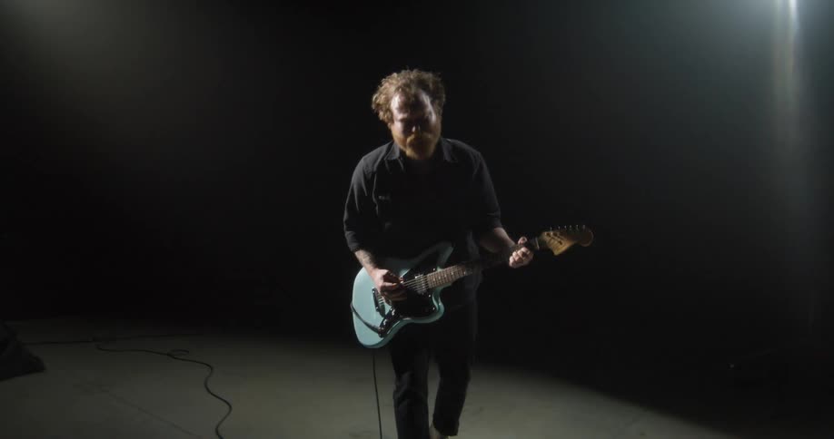 A man with a beard and messy hair plays a light blue electric guitar in a dark room with a bright spotlight