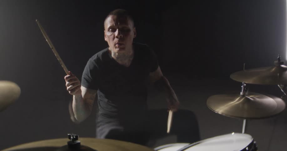 An energetic close view shows a tattooed drummer in a black tshirt playing a drum set in a dark room with bright lights