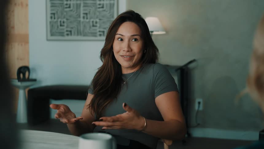 A woman with long brown hair sits at a table and talks to someone off camera