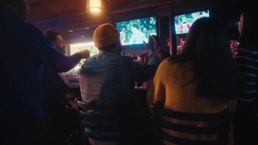 A group of friends sits at a dimly lit bar cheering and gesturing while watching a soccer match on multiple wall mounted screens