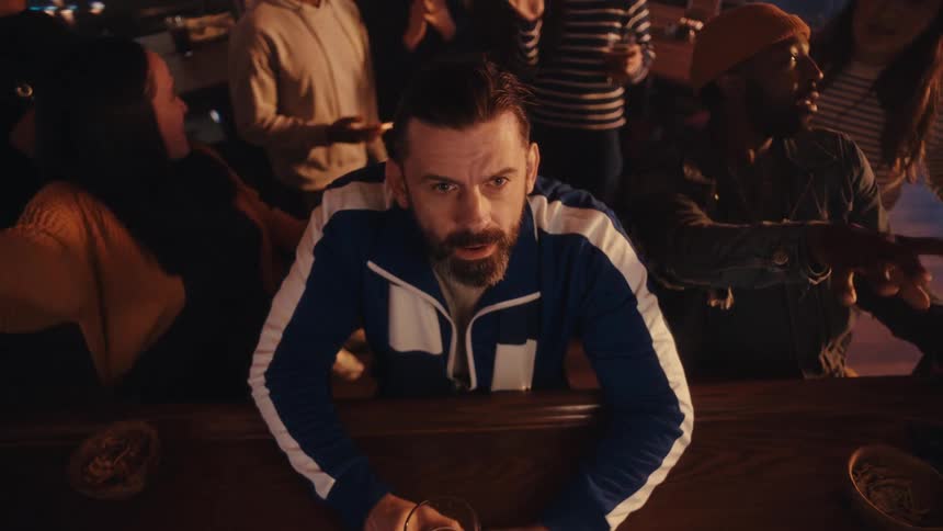 Man seated at a bar counter holds a glass while people socialize around him