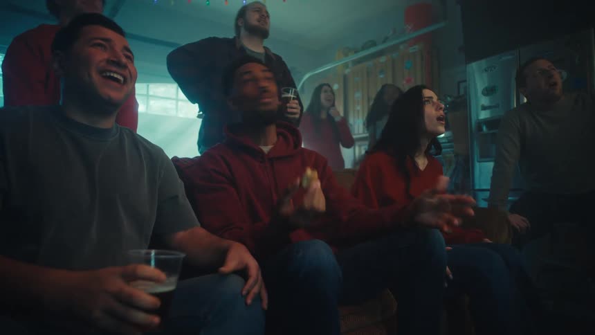 Group of people gathered in a living room watching something offscreen with varied reactions during a social gathering