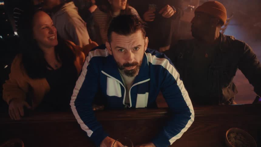 A man with a beard and a blue track jacket leans on a wooden bar while several people behind him cheer and gesture excitedly