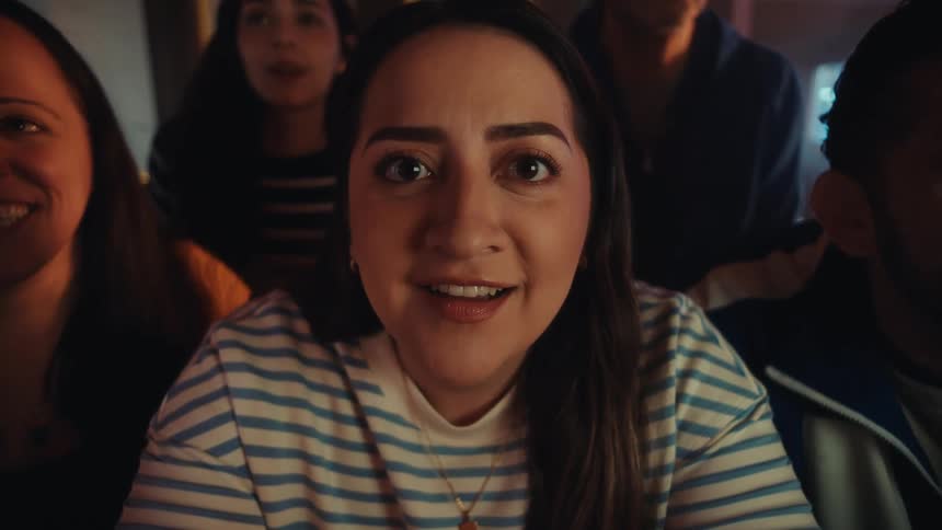 People standing in a room as light from a television shines on a young womans face and they cheer in excitement