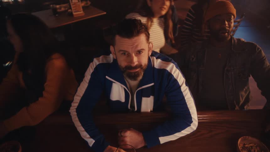 A man in a blue and white tracksuit leans against a bar counter in a crowded room and looks directly at the camera